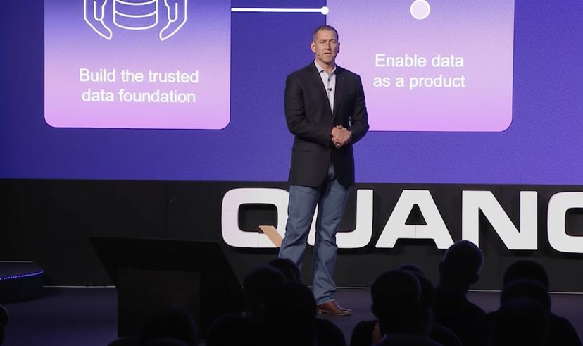 Man in a dark blazer talks on stage, standing in front of a screen displaying tech-related text and diagrams.
