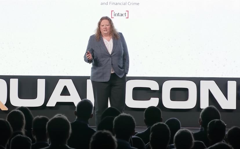A person speaks on stage at a conference, standing before an audience. The background features a large screen displaying part of the word "QUANCON."