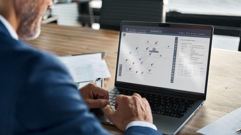 Person in a suit using a laptop displaying a complex network diagram on the screen, sitting at a wooden desk.