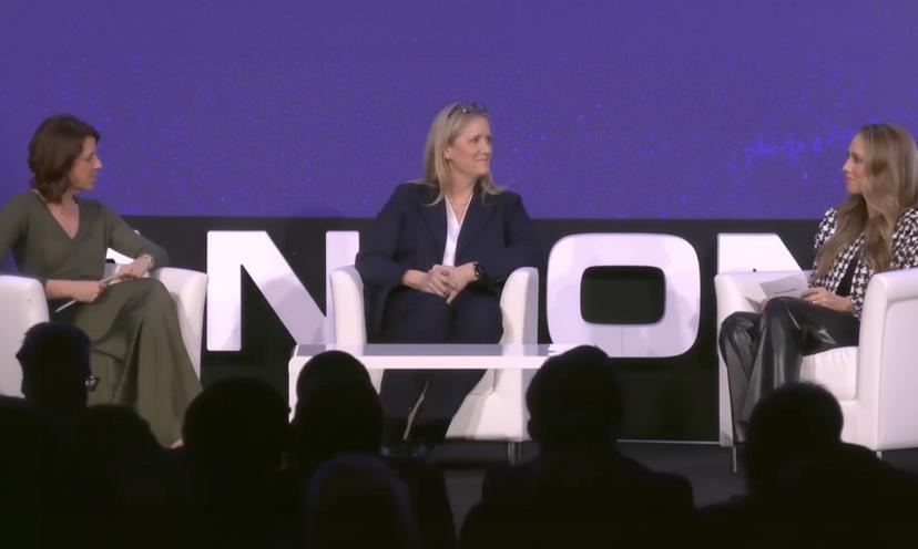 Three women seated on a stage engaged in a panel discussion, with a large screen displaying text behind them.