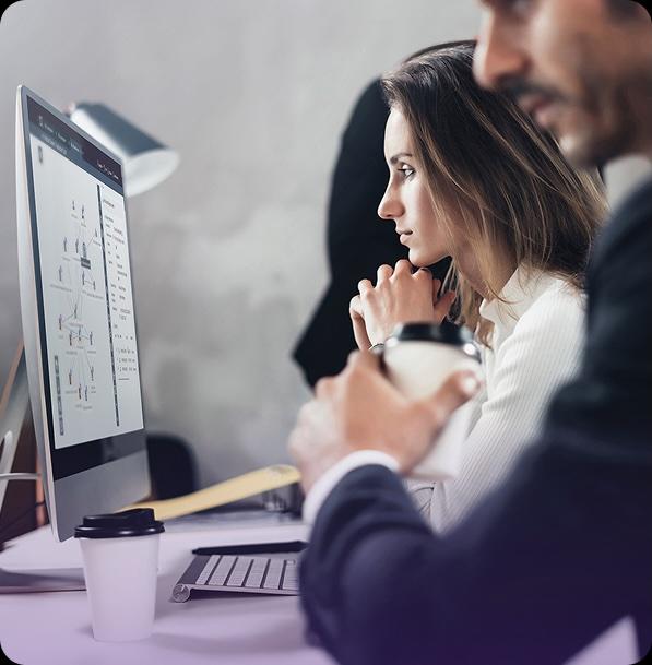 Two people working at computers in a modern office, one focusing on a screen displaying diagrams, the other holding a coffee cup.