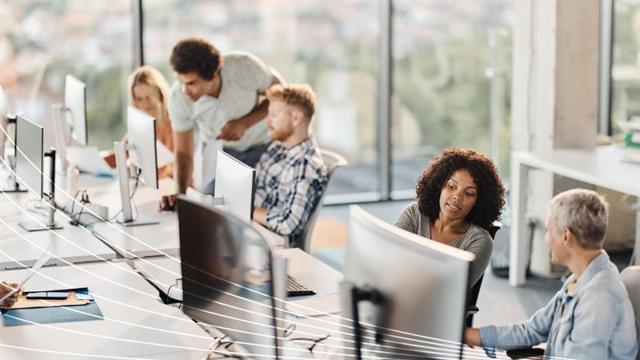 A diverse group of people working and collaborating at computer desks in a modern office with large windows in the background.