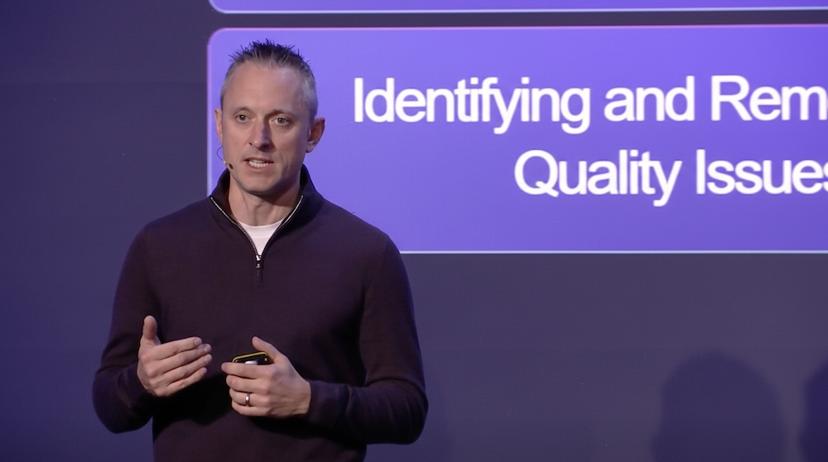 Jamie Hutton speaking on stage about quality issues, standing in front of a presentation slide with a microphone on his cheek.