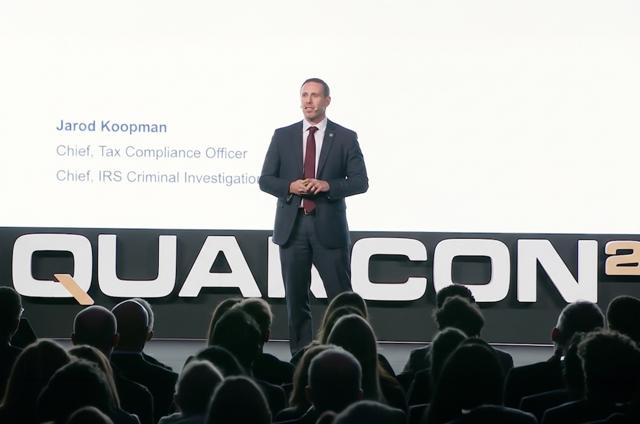 A man in a suit speaks on stage at QUANCON26. The screen behind displays his roles, including IRS Chief of Tax Compliance. Audience silhouettes visible.