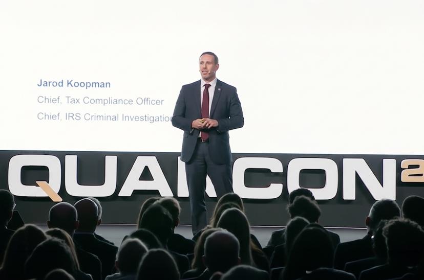 A man in a suit speaks on stage at QUANCON26. The screen behind displays his roles, including IRS Chief of Tax Compliance. Audience silhouettes visible.