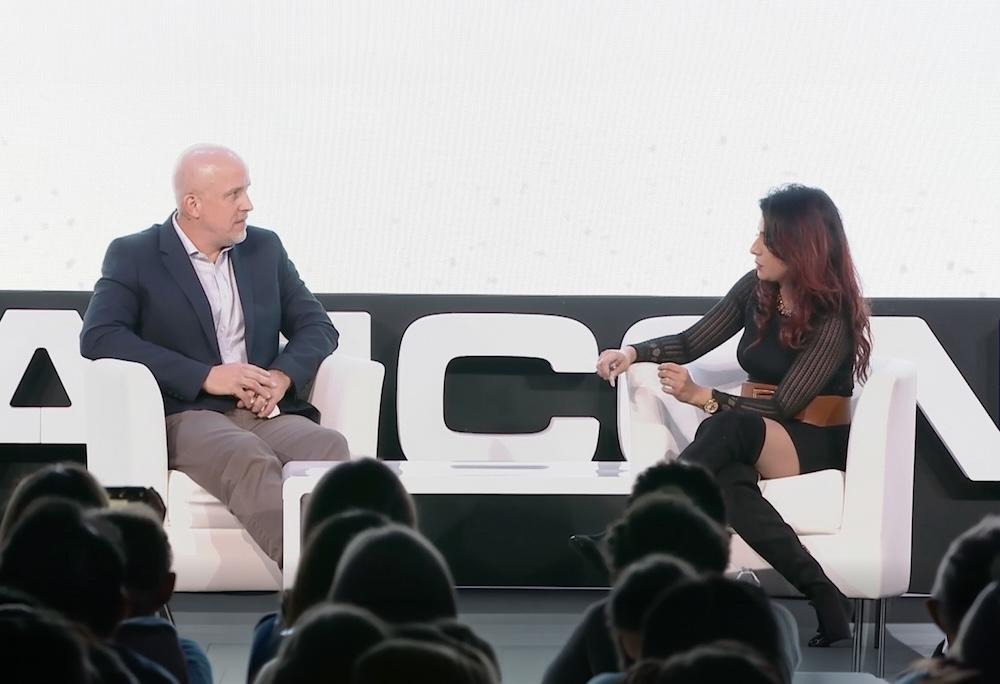 Two people seated on stage with a white backdrop, engaged in conversation. An audience is visible in the foreground.