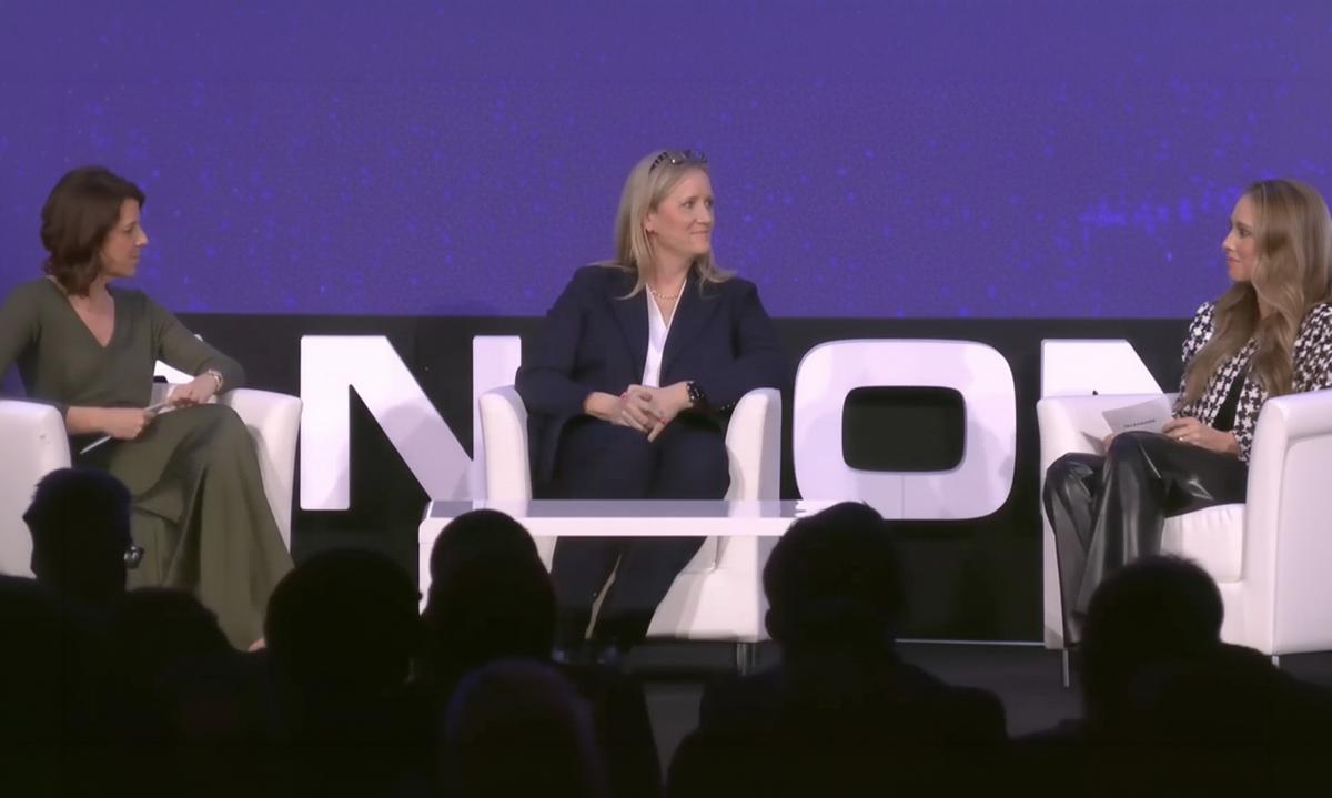Three women seated on a stage engaged in a panel discussion, with a large screen displaying text behind them.