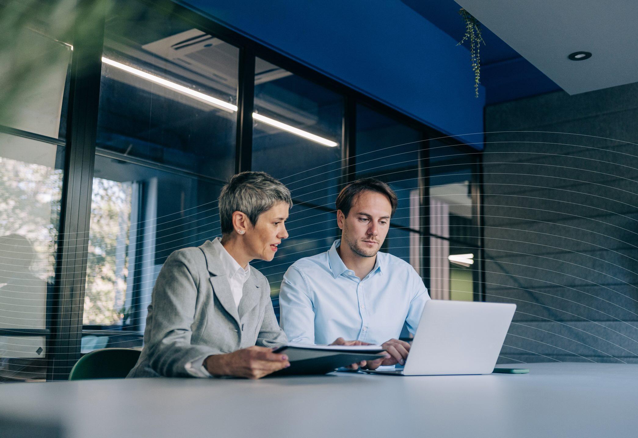 Two people in business attire sit at a table with a laptop, engaged in discussion. A large window and modern office decor are in the background.