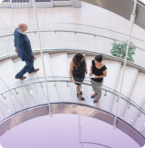Two women stand on a curved staircase, conversing, while a man in a suit walks past. The setting is a modern, open indoor space.