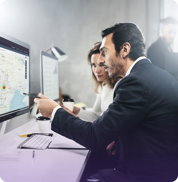 Man and woman in an office discussing a map on a computer screen, with a notebook and coffee cups on the desk.